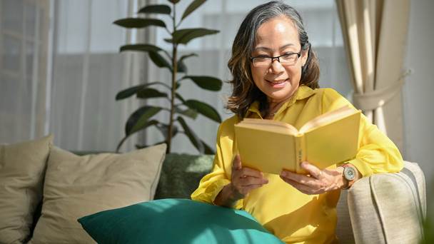 Mulher grisalha sentada em sofá, lendo um livro, relaxada, em um ambiente acolhedor, com luz natural.